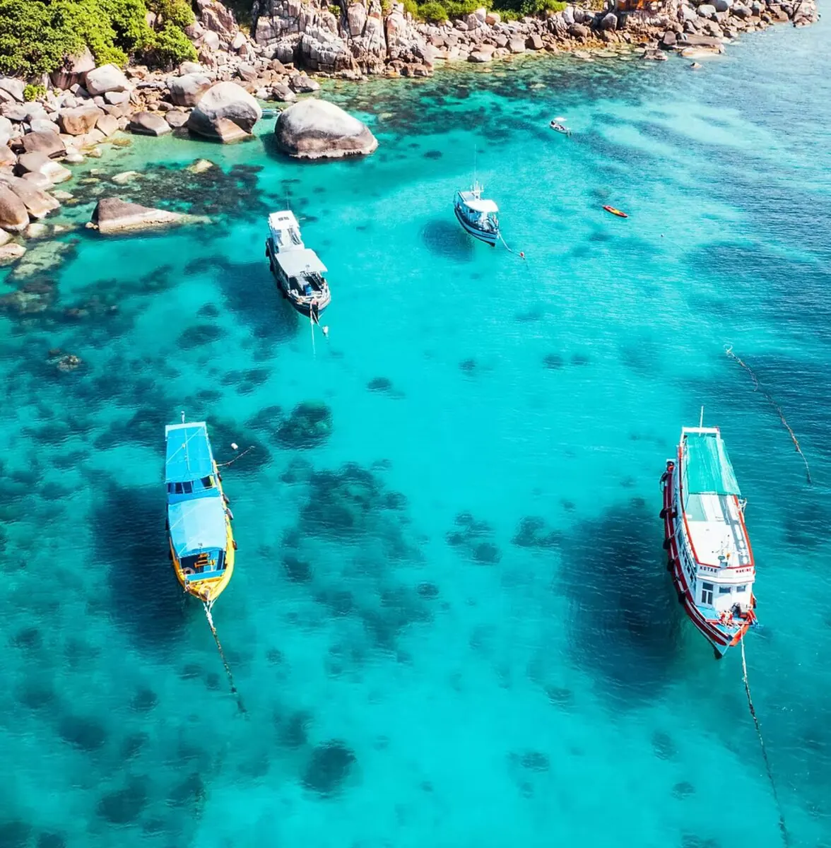 Aerial view of long-tail boats over crystal-clear water at a Koh Tao bay, granite boulders breaking the turquoise