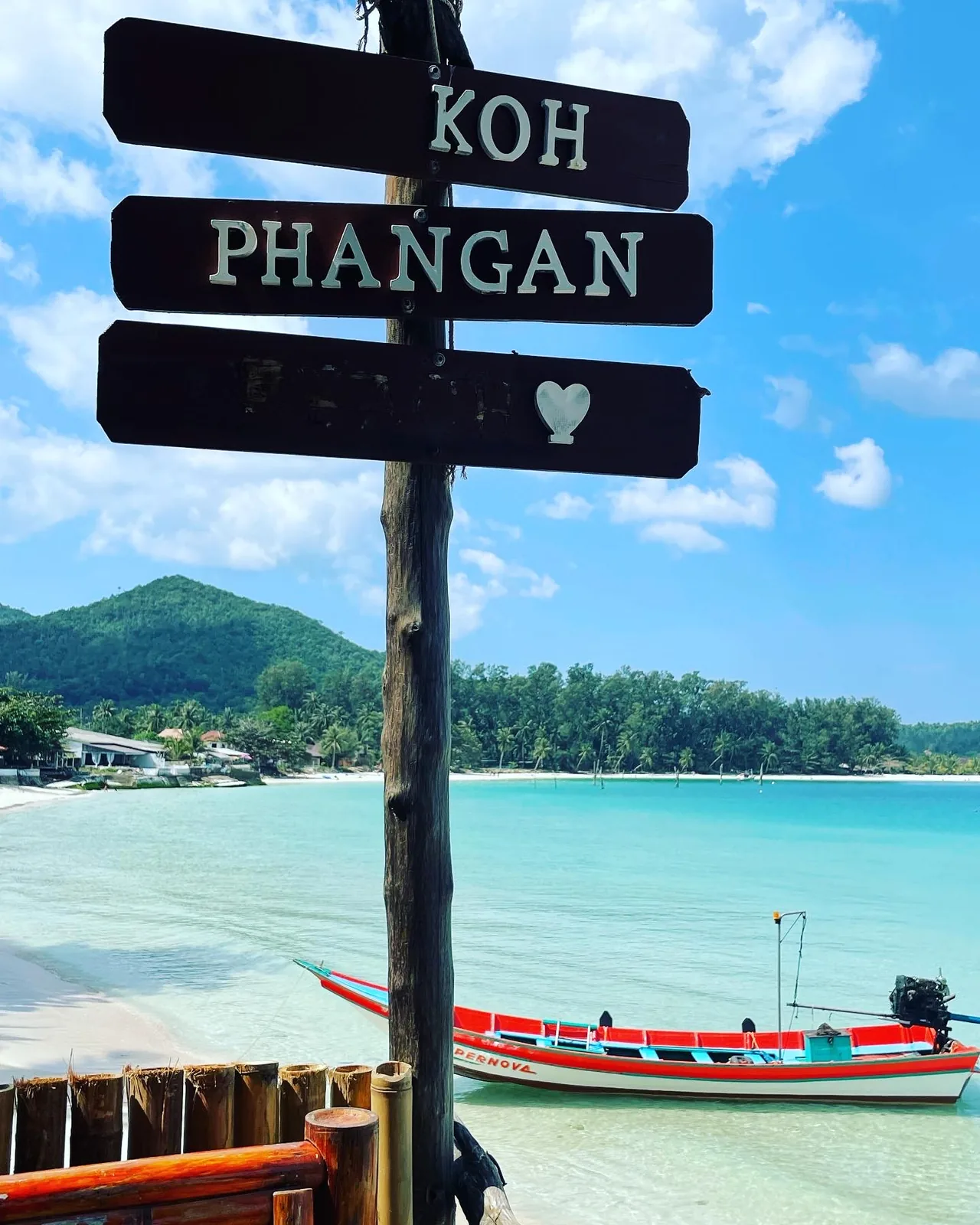 A Brown sign with white writing Koh Phangan near the beach with a long tail boat