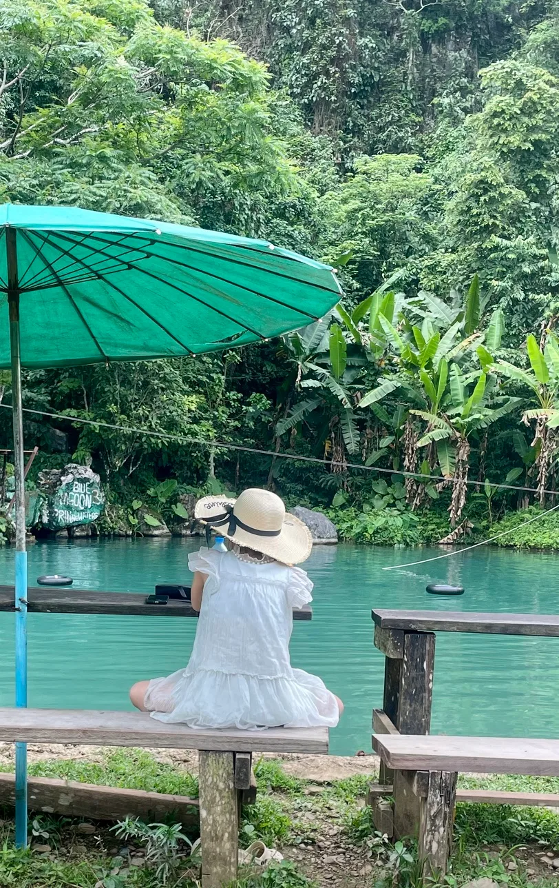 Blue Lagoon number 3 in Laos and a woman in white with a sun hat sitting under a green umbrella looking at the blue water